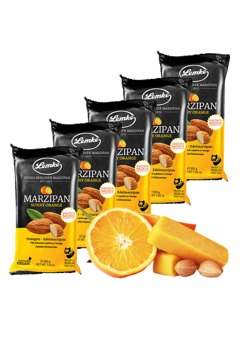 Five Lemke Sunny Orange marzipan packs arranged behind orange slices. Almonds and orange marzipan bars sit in front to show the almond and citrus flavors.
