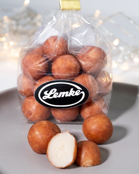 Clear Lemke bag filled with round marzipan potatoes on a grey surface. One cut piece in front shows the pale marzipan center inside the golden brown cocoa coating.