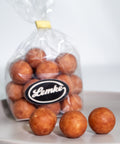 Clear Lemke bag filled with round cocoa dusted marzipan potatoes on a grey surface. Three marzipan balls sit in front to show the golden brown coating and rounded shape.