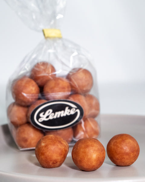 Clear Lemke bag filled with round cocoa dusted marzipan potatoes on a grey surface. Three marzipan balls sit in front to show the golden brown coating and rounded shape.