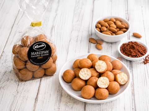 Lemke Marzipan Kartoffel-Konfekt Zimt bag beside a plate of cinnamon marzipan potato sweets, almonds, and cocoa powder on a white wooden tabletop.